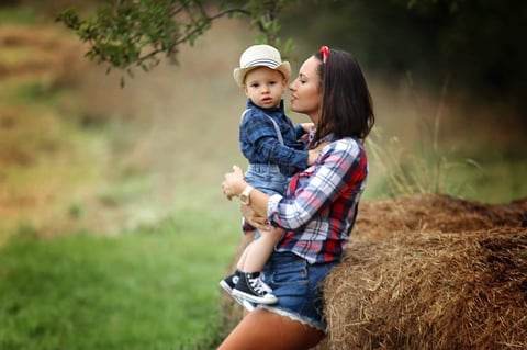 Woman holding toddler boy wearing white hat sitting on hay bale in outdoor setting