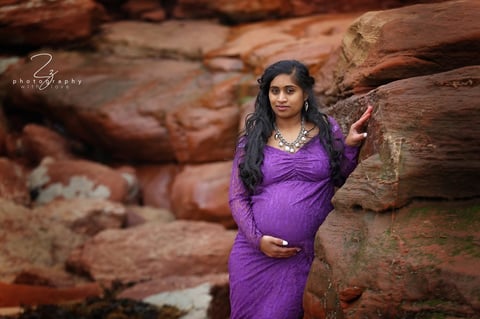 Pregnant woman in purple dress posing against rustic wooden logs outdoors