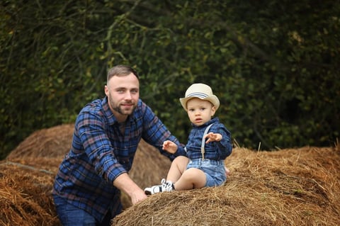 Man and toddler in denim shirts sitting on hay bales with green foliage background
