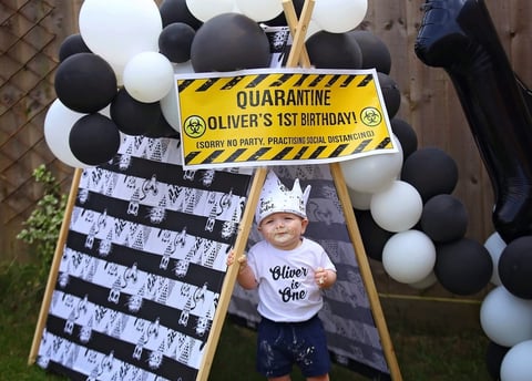 Quarantine-themed first birthday display with yellow caution sign, black and white balloons, and photo of child wearing Oliver is One shirt