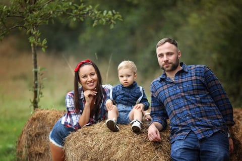 Family of three sitting on a hay bale outdoors with green foliage background